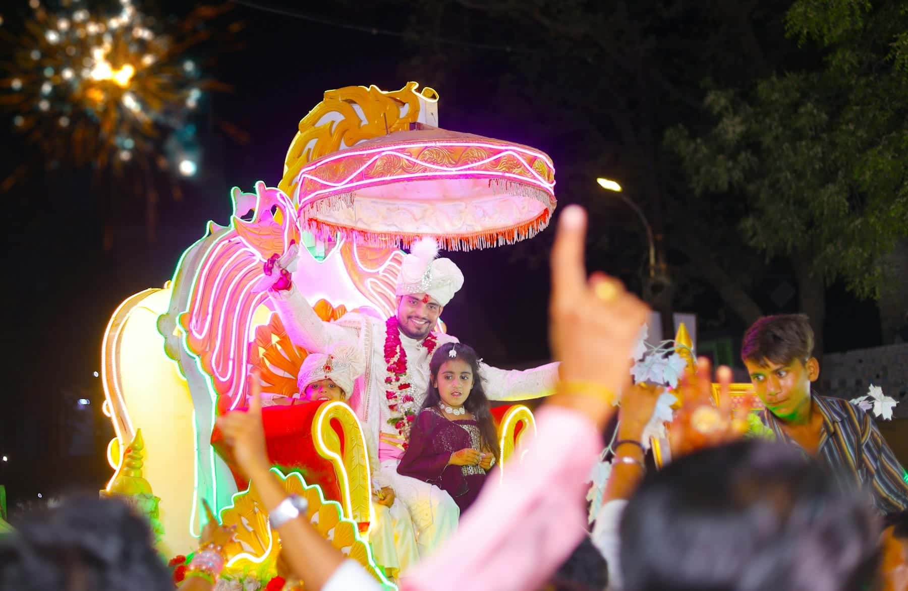 Groom seated on an illuminated baraat float as guests dance and cheer around him at night