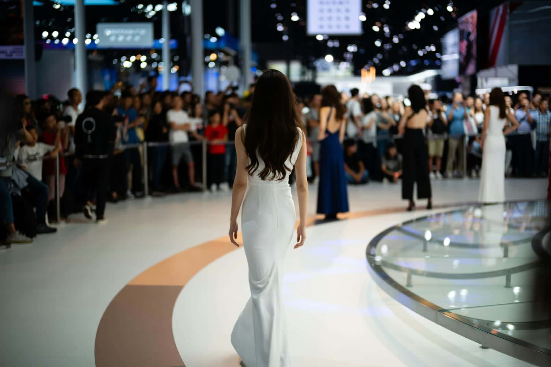 Models in evening gowns walking a curved runway past a crowd at an exhibition