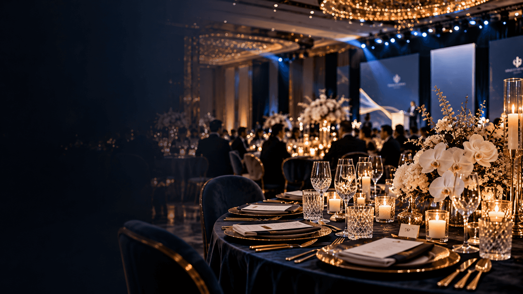 Gala dinner table laid with gold chargers, crystal glasses, white orchids and candles, a lit stage behind