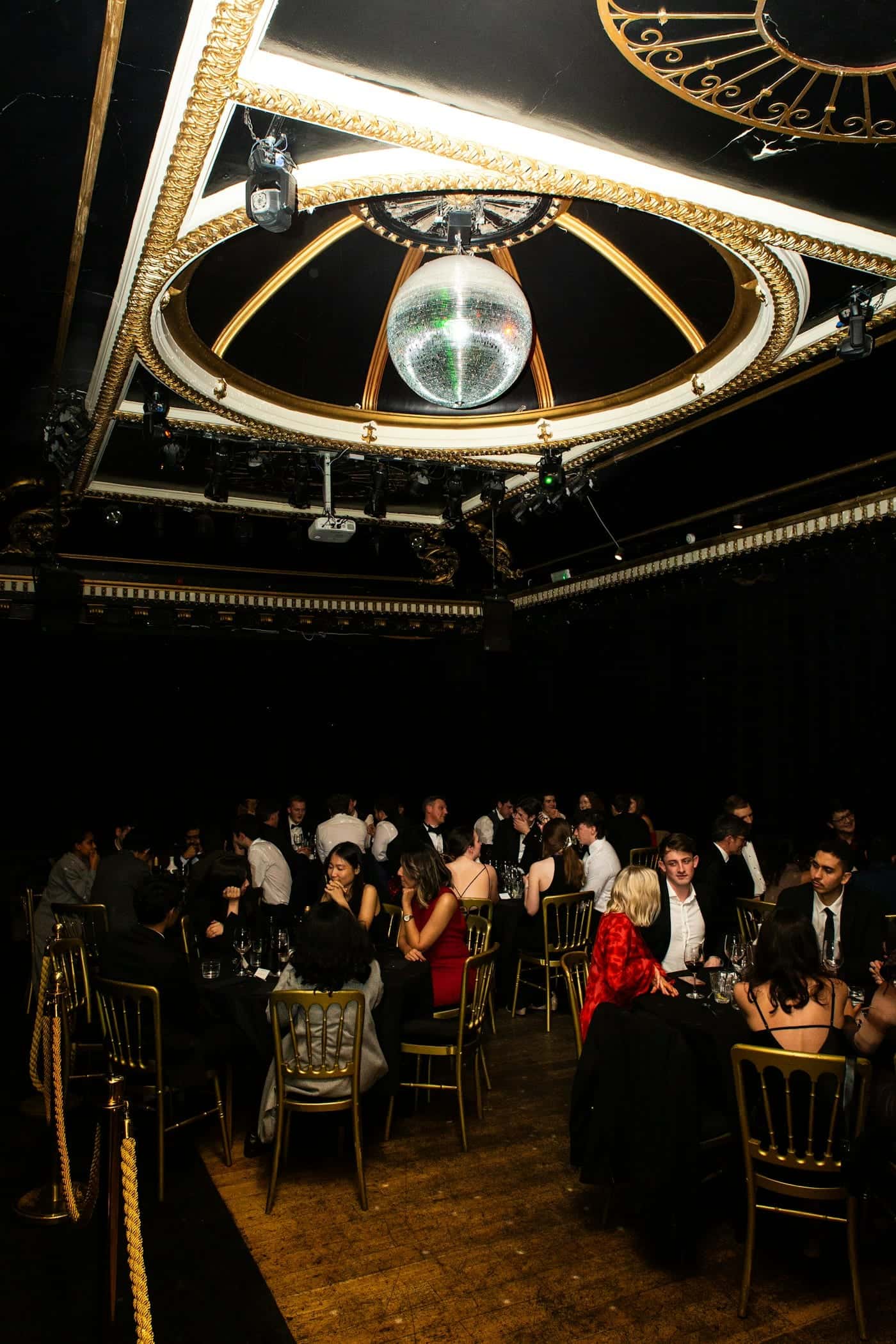Guests in evening wear seated at round tables in a dark hall beneath a mirror ball