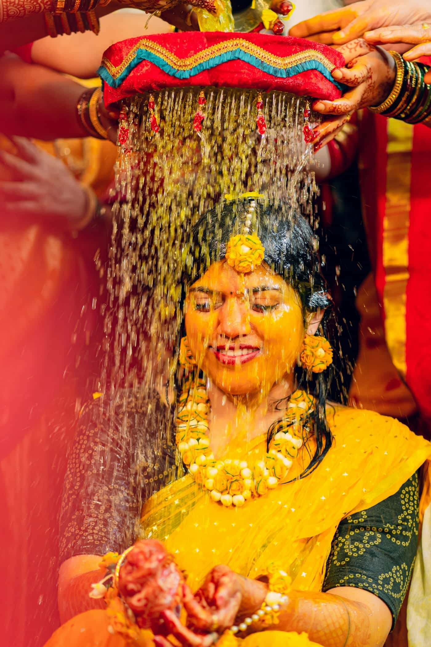 Bride in a yellow sari laughing as family shower her with flowers at a haldi ceremony