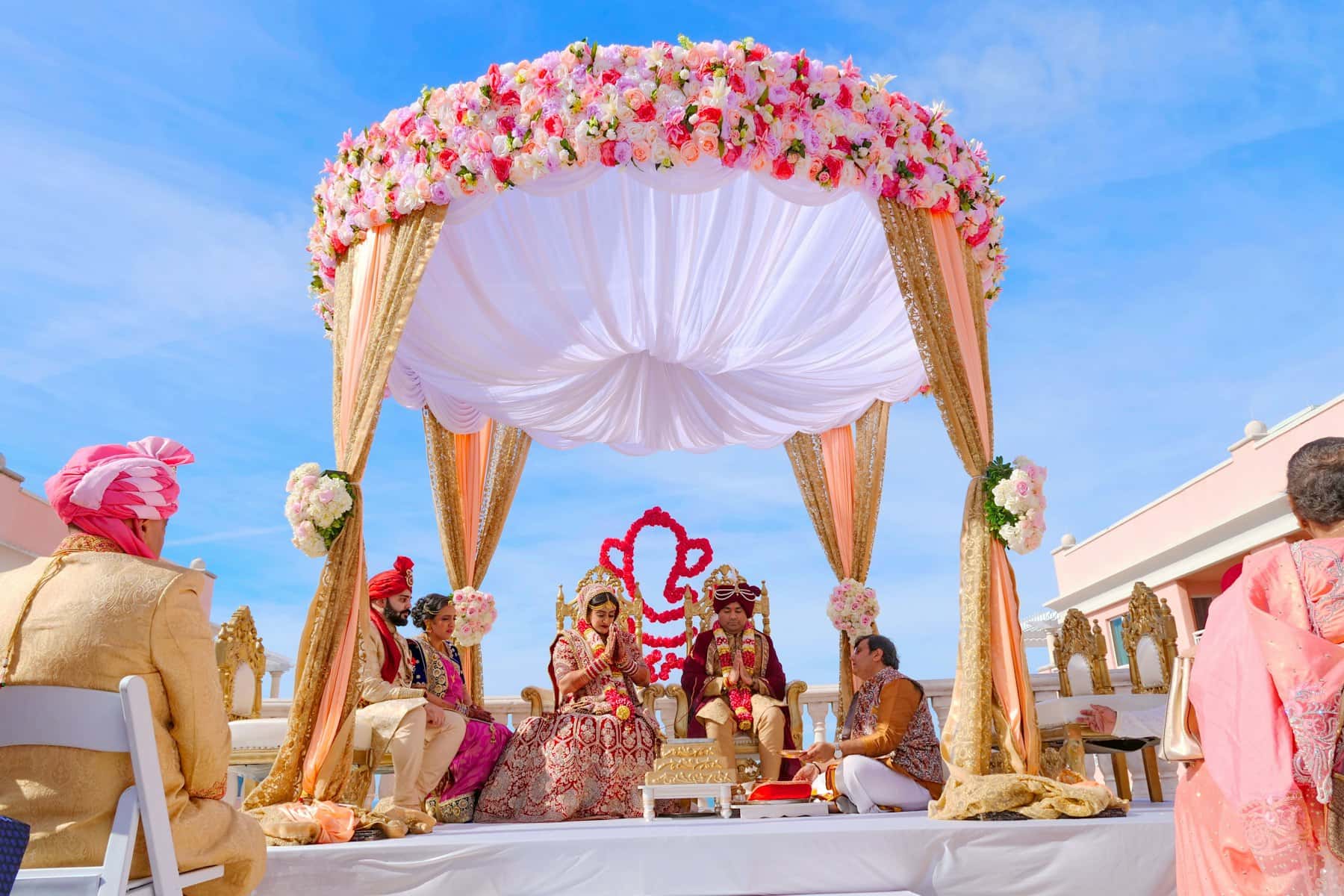 Couple in traditional wedding attire beneath a pink and white floral mandap draped in gold fabric, family seated beside them