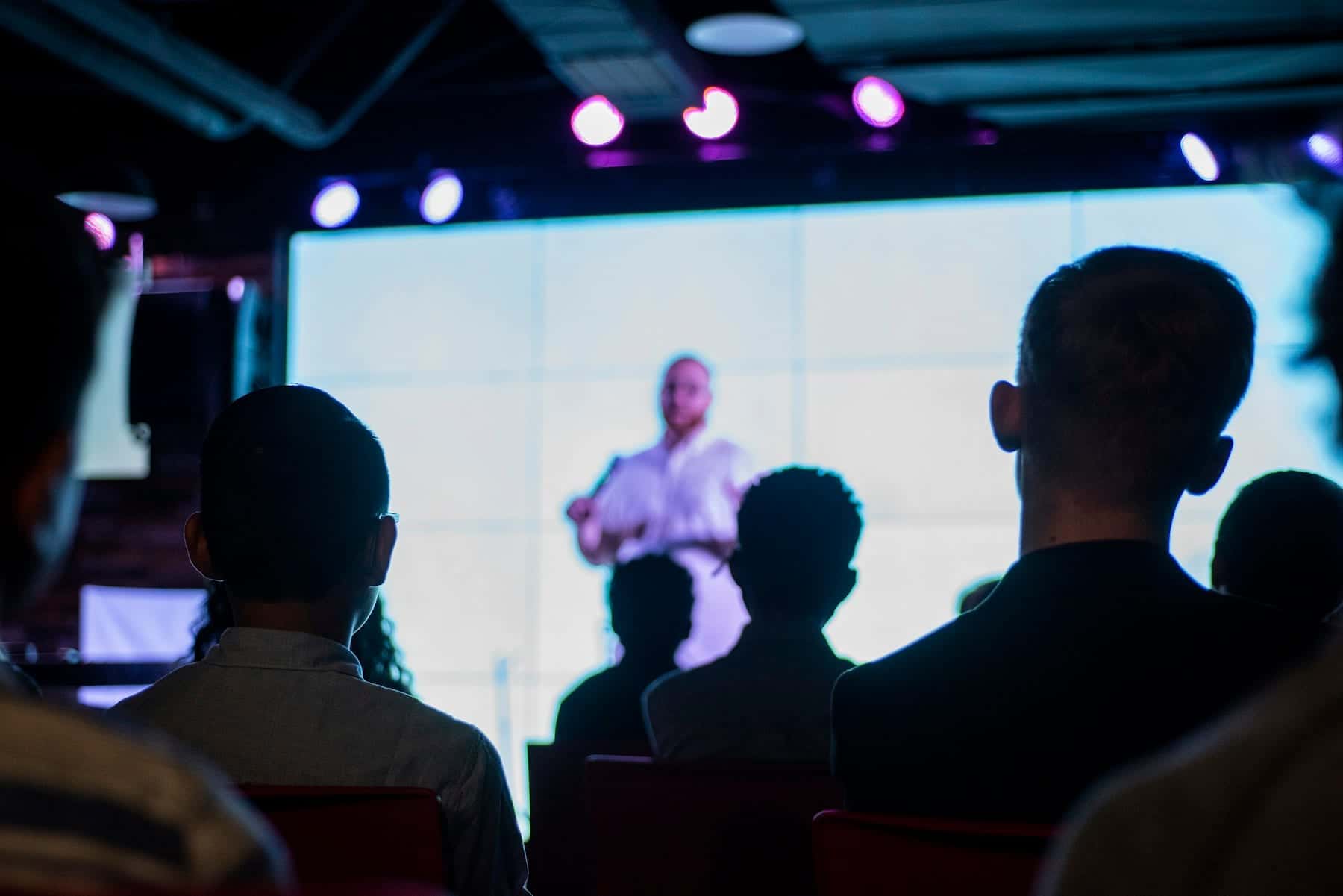 Audience in silhouette watching a speaker projected on a large screen at a conference