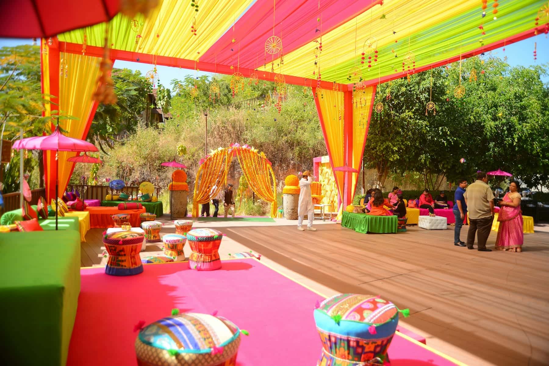 Outdoor mehendi set-up with pink and yellow canopies, colourful floor cushions and a floral archway