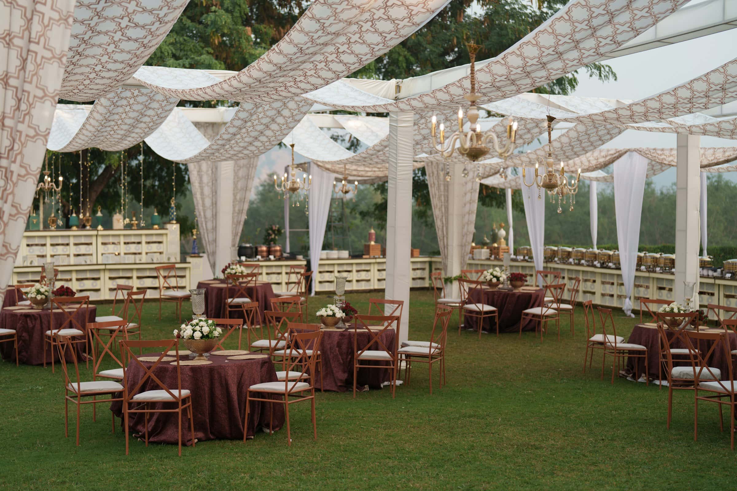 Lawn reception under draped patterned canopies and chandeliers, with round tables set for dinner