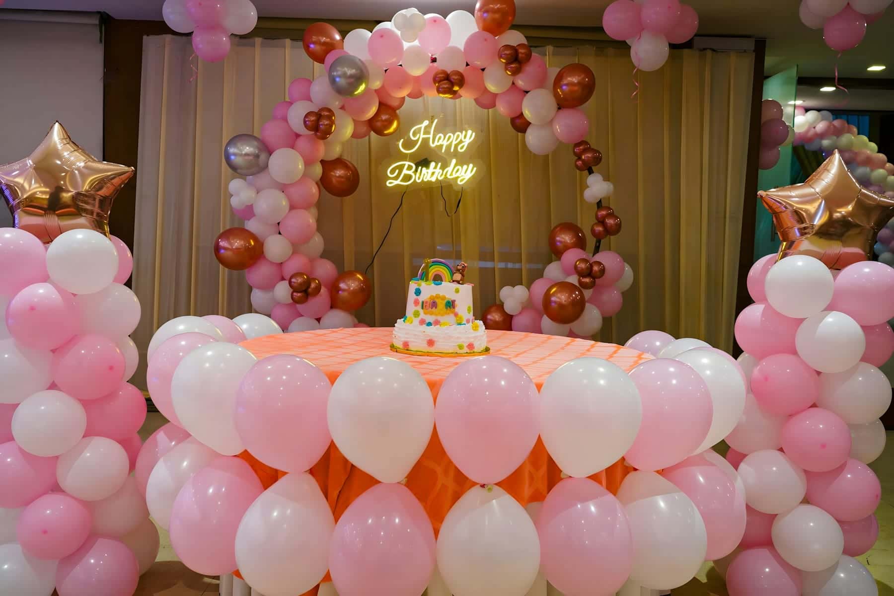 Pink and white balloon arch with a neon Happy Birthday sign behind a rainbow birthday cake