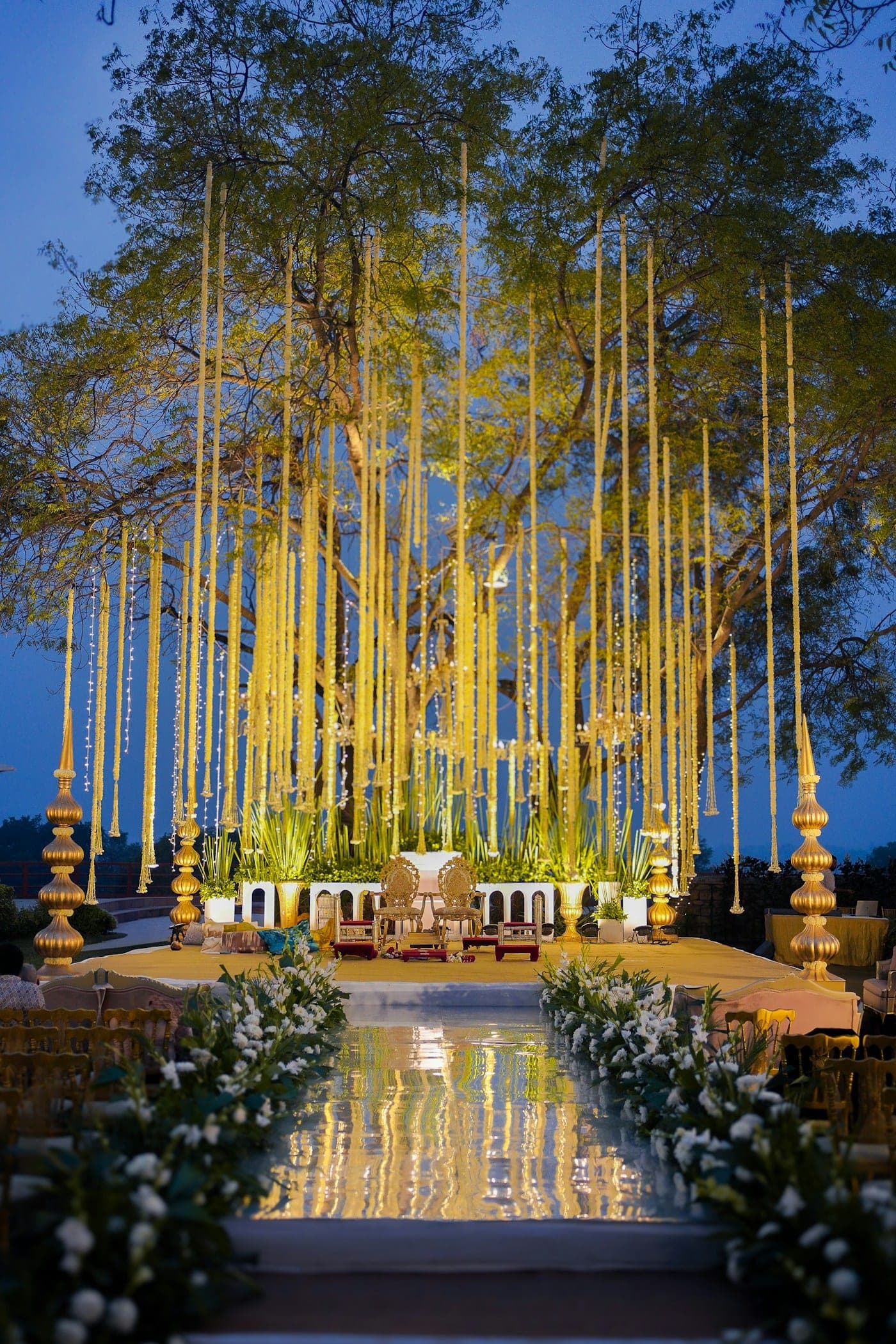Wedding stage at dusk beneath a canopy of glowing hanging lights, with a white floral aisle leading to it