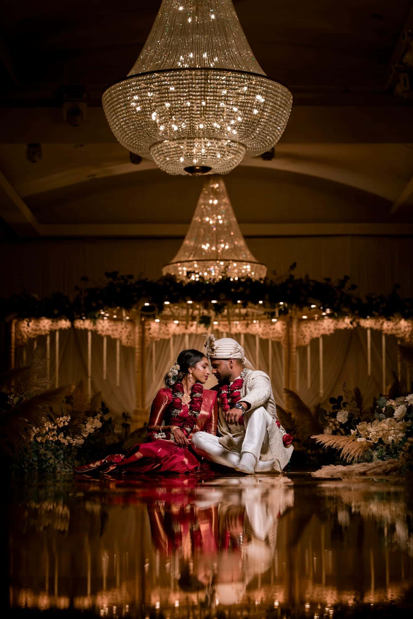 Bride and groom seated together beneath crystal chandeliers, reflected in a glossy floor