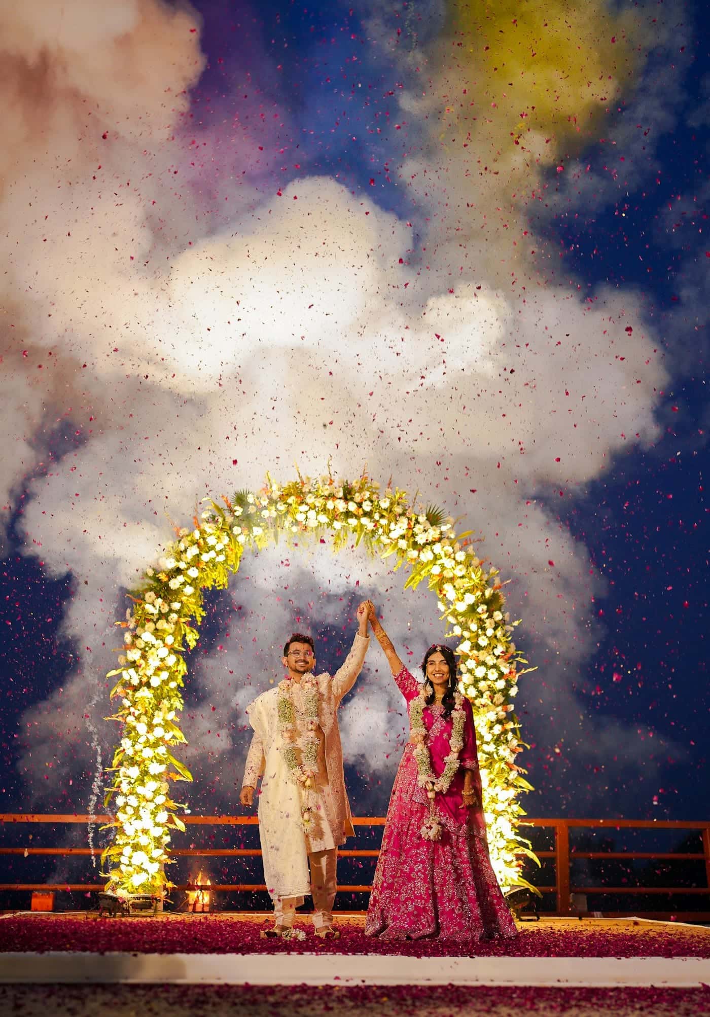 Couple raising their joined hands beneath an illuminated floral arch as confetti rises around them