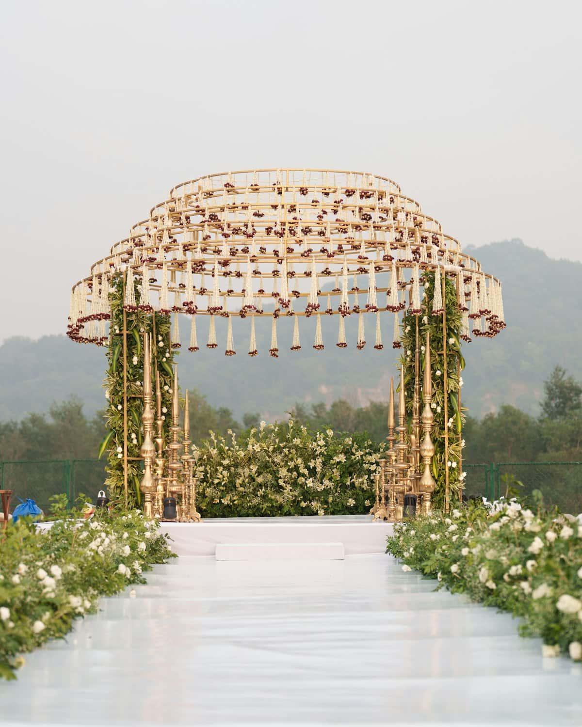 White flower-lined aisle leading to a gold mandap hung with jasmine strands, hills behind