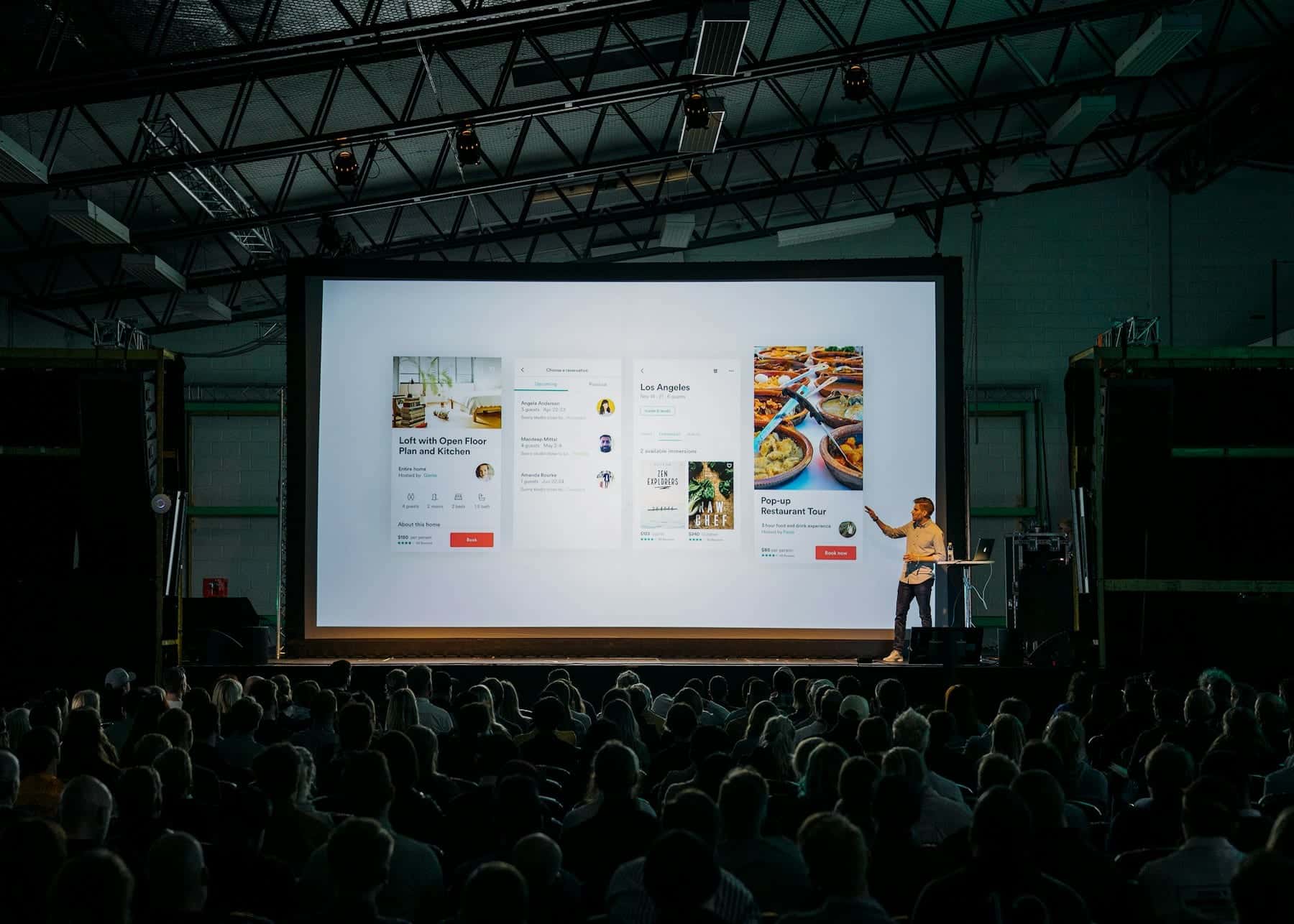 Presenter beside a giant screen showing app designs, facing a packed audience in a warehouse venue