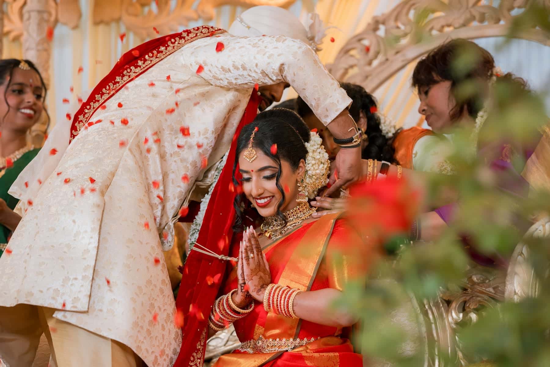 Groom leaning towards the bride as guests shower the couple with rose petals during a Hindu wedding ceremony